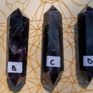 May include: Three black amethyst crystal points with labels "B", "C", and "D" arranged on a wooden board with a geometric pattern.