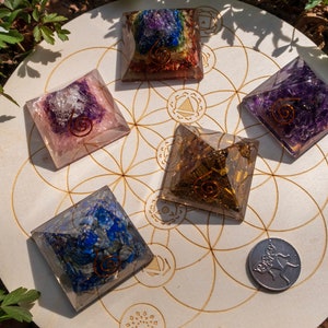 May include: Six crystal pyramids of various colours, including amethyst, rose quartz, and lapis lazuli, are arranged on a wooden circle with a flower of life design. The pyramids are all approximately 5 cm in size and have a copper wire spiral inside.