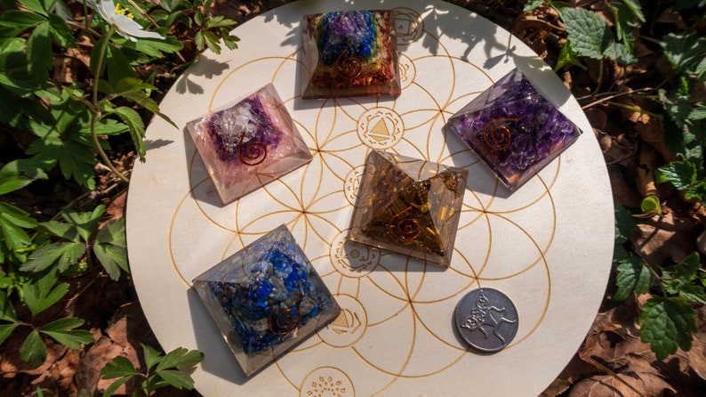 May include: Six crystal pyramids of various colours, including amethyst, rose quartz, and lapis lazuli, are arranged on a wooden circle with a flower of life design. The pyramids are all approximately 5 cm in size and have a copper wire spiral inside.