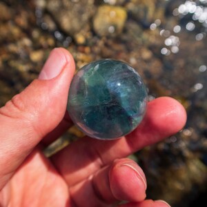 Puede incluir: Una esfera de fluorita verde y azul, sostenida en una mano, con un fondo borroso de rocas y agua.