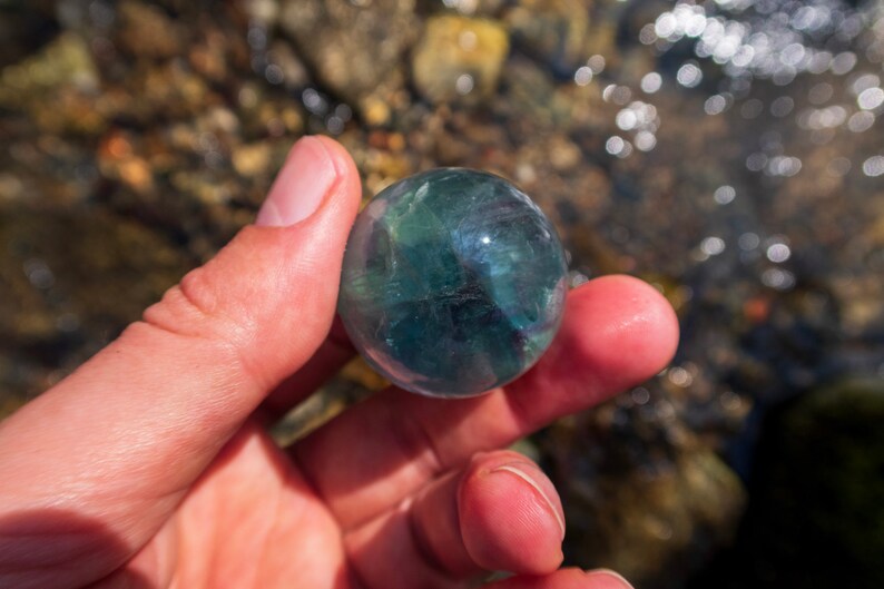 Puede incluir: Una esfera de fluorita verde y azul, sostenida en una mano, con un fondo borroso de rocas y agua.