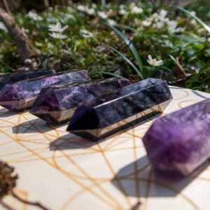 May include: Five amethyst crystal wands, with a purple and black color, are arranged on a wooden surface with a geometric pattern. The wands are pointed at both ends and have a smooth, polished finish.