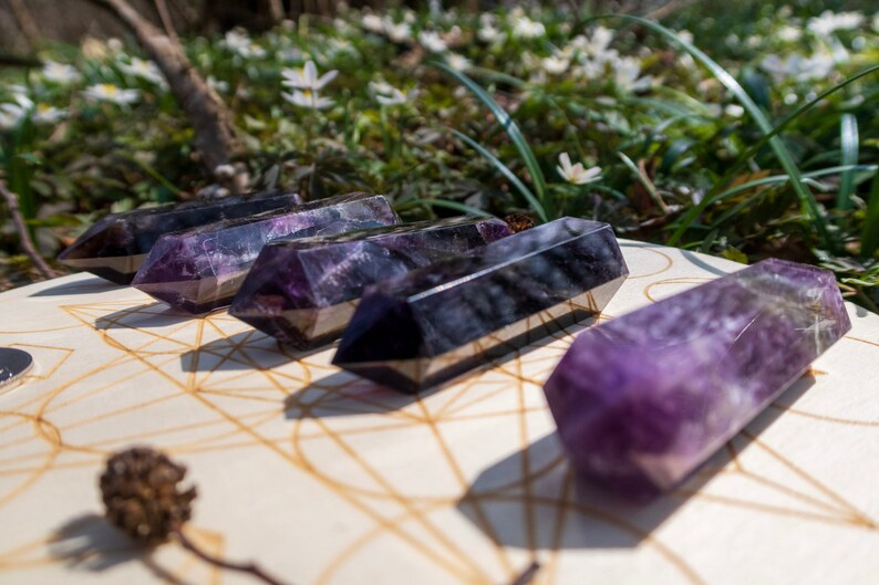 May include: Five amethyst crystal wands, with a purple and black color, are arranged on a wooden surface with a geometric pattern. The wands are pointed at both ends and have a smooth, polished finish.
