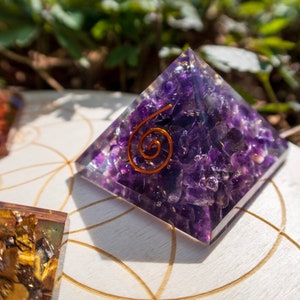 May include: A purple amethyst crystal pyramid with a copper wire spiral on top. The pyramid is sitting on a white wooden surface with a geometric pattern.