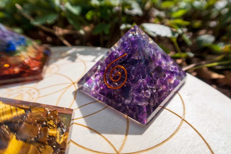 May include: A purple amethyst crystal pyramid with a copper wire spiral on top. The pyramid is sitting on a white wooden surface with a geometric pattern.