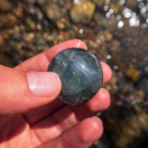 Puede incluir: Una piedra redonda, lisa, de color verde oscuro y negro con una superficie brillante. La piedra est&aacute; siendo sostenida en una mano.