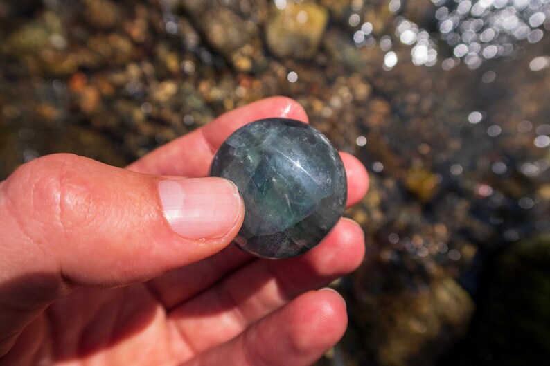 Puede incluir: Una piedra redonda, lisa, de color verde oscuro y negro con una superficie brillante. La piedra est&aacute; siendo sostenida en una mano.