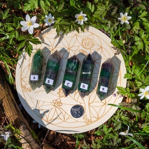 May include: Five green and purple fluorite points arranged in a row on a wooden circle with a flower of life design. The points are labeled A through E.