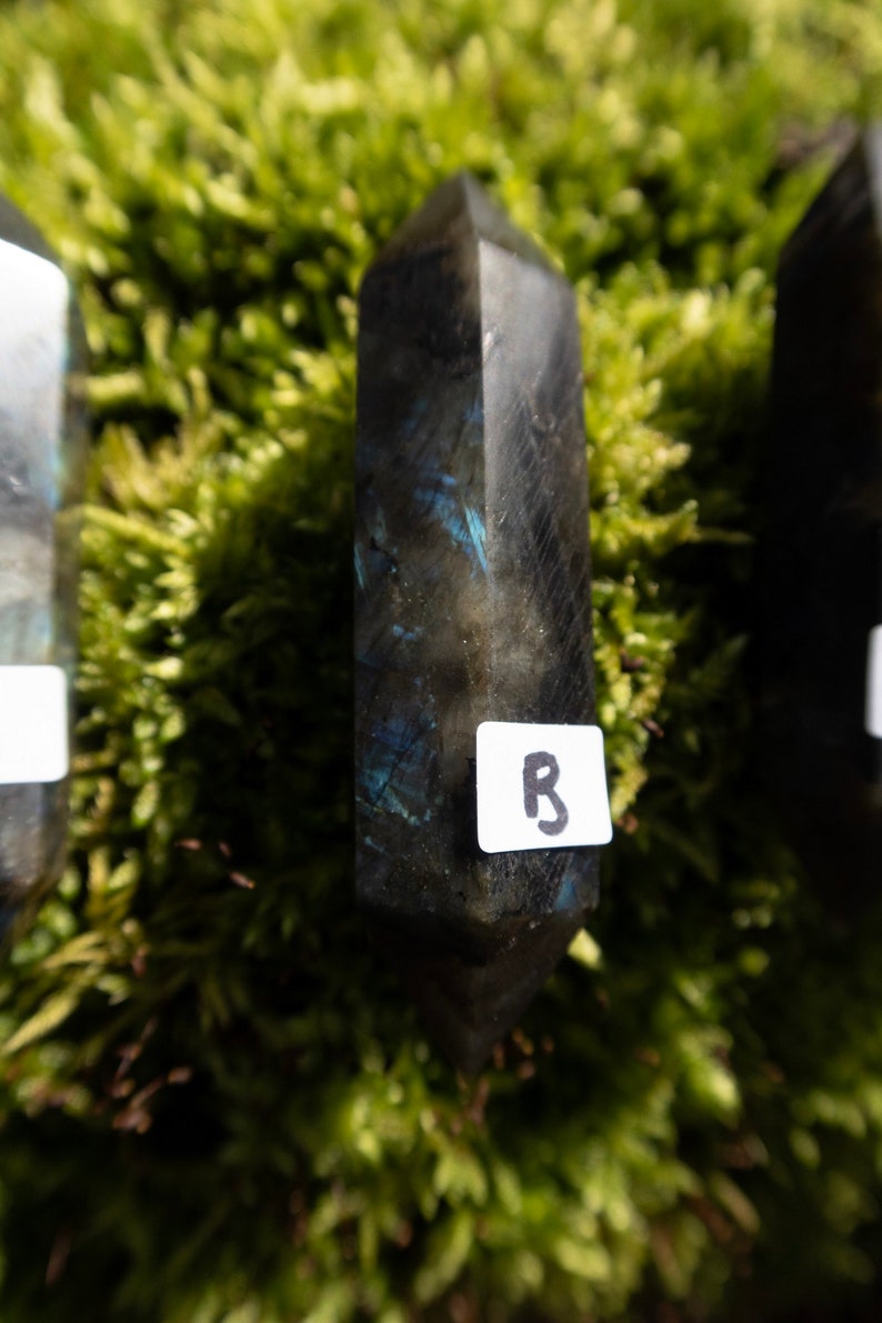 Op de afbeelding: Een donkergrijze labradorit-kristalpunt met blauwe, iriserende flitsen. Het kristal is gemarkeerd met de letter "B".