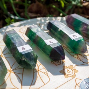 May include: Five green and purple fluorite crystal points labeled A through E, arranged on a white wooden surface with a circular design.