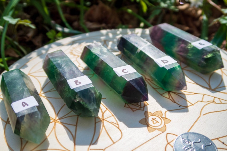 May include: Five green and purple fluorite crystal points labeled A through E, arranged on a white wooden surface with a circular design.