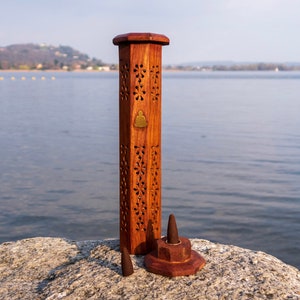 May include: A wooden incense burner with intricate carvings and a small brass Buddha design. The burner is standing on a rock near a body of water.