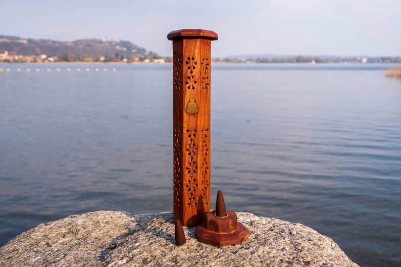 May include: A wooden incense burner with intricate carvings and a small brass Buddha design. The burner is standing on a rock near a body of water.