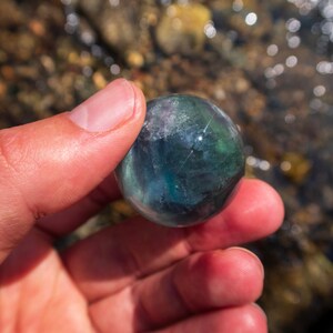 Puede incluir: Una esfera de fluorita verde y azul, un tipo de cristal, se sostiene en una mano. La esfera es lisa y pulida.