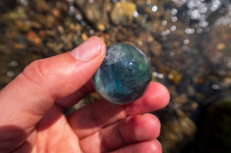 Puede incluir: Una esfera de fluorita verde y azul, un tipo de cristal, se sostiene en una mano. La esfera es lisa y pulida.