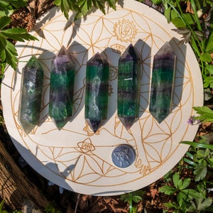 May include: Six green, purple, and white fluorite crystal points arranged in a row on a wooden circle with a flower of life design.