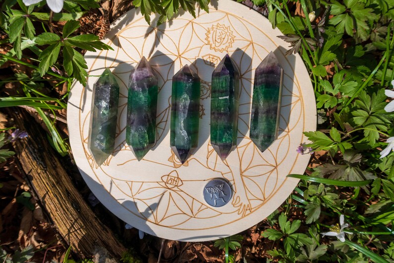 May include: Six green, purple, and white fluorite crystal points arranged in a row on a wooden circle with a flower of life design.