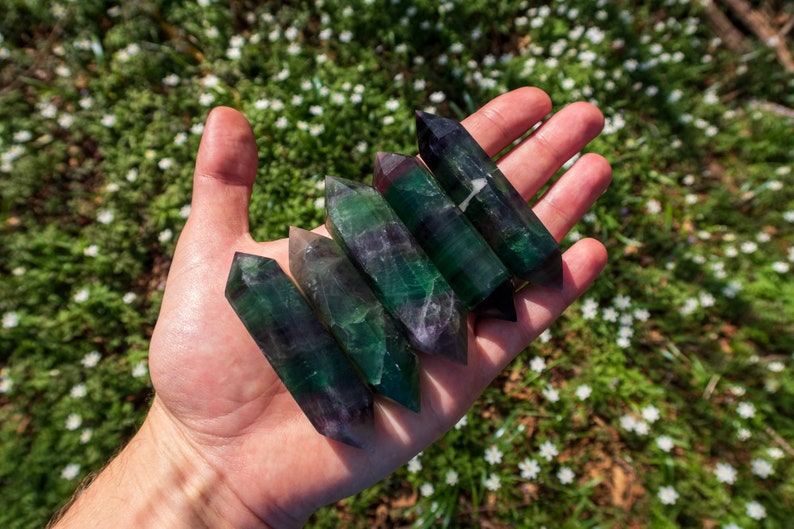 May include: A hand holding six green, purple, and black fluorite crystal points. The points are smooth and polished, and they have a variety of shapes and sizes.