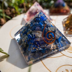 May include: A clear crystal pyramid filled with blue and white stones, with a copper wire spiral design. The pyramid is sitting on a white wooden surface with a circular design.