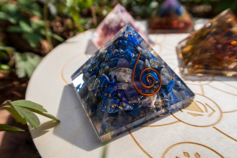 May include: A clear crystal pyramid filled with blue and white stones, with a copper wire spiral design. The pyramid is sitting on a white wooden surface with a circular design.