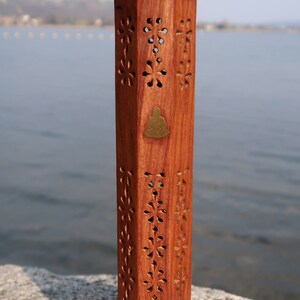 May include: A wooden incense burner with intricate carvings in the shape of flowers. The burner is a hexagonal shape and has a brass Buddha figure on the front. The burner is sitting on a rock.