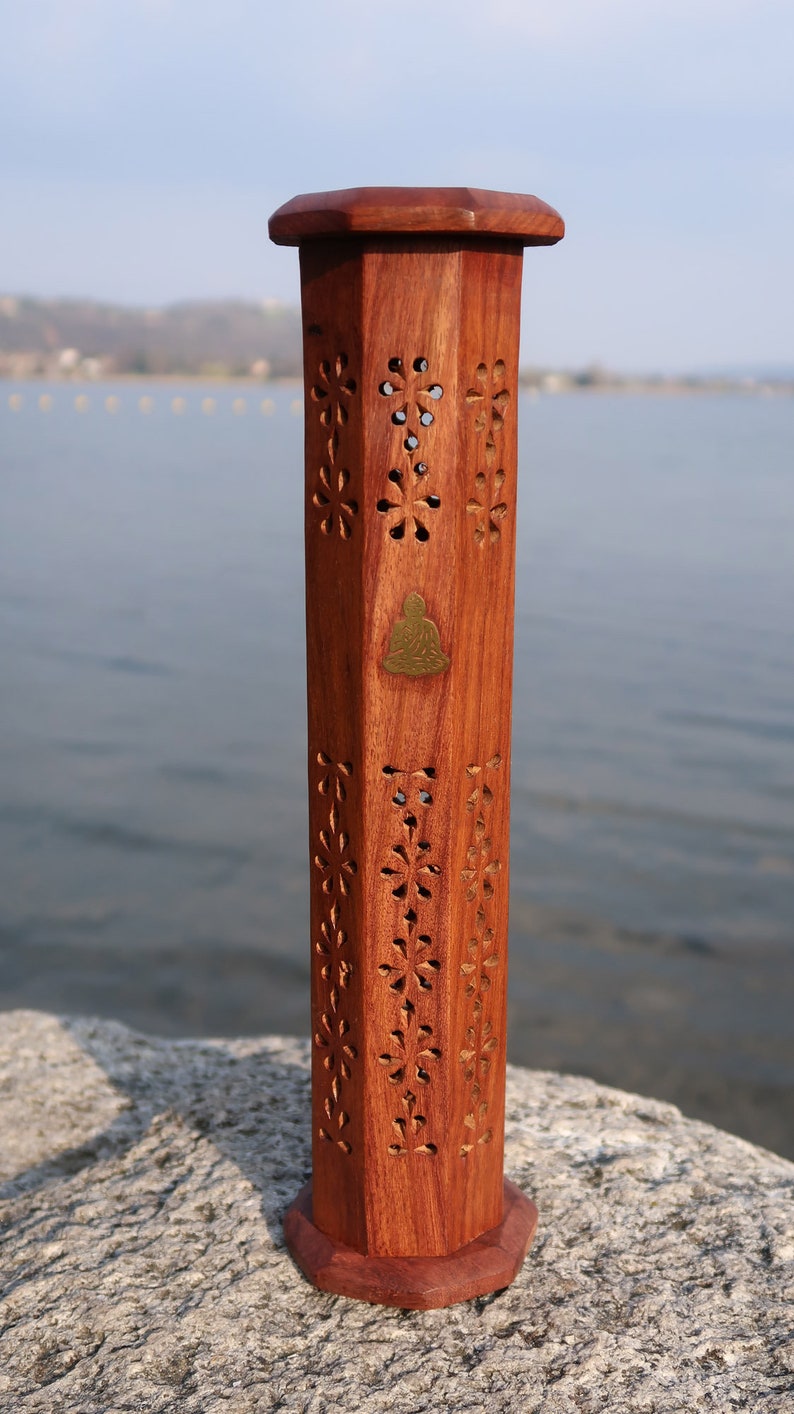 May include: A wooden incense burner with intricate carvings in the shape of flowers. The burner is a hexagonal shape and has a brass Buddha figure on the front. The burner is sitting on a rock.