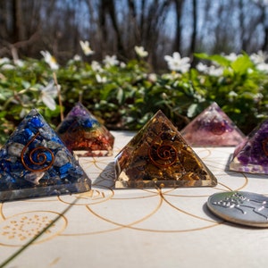May include: Five crystal pyramids with different coloured stones, including blue, gold, pink, and purple, are arranged on a wooden circle with a silver coin in the centre. The pyramids are made of clear resin with a copper wire spiral inside.