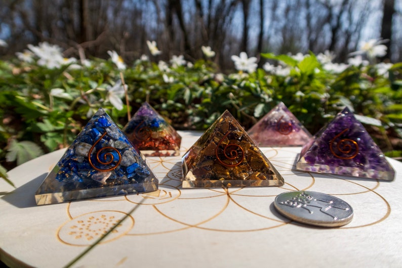 May include: Five crystal pyramids with different coloured stones, including blue, gold, pink, and purple, are arranged on a wooden circle with a silver coin in the centre. The pyramids are made of clear resin with a copper wire spiral inside.