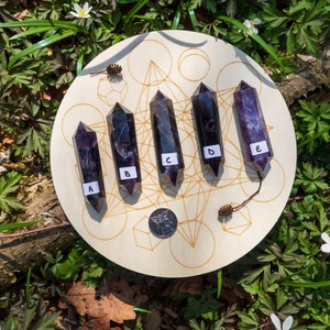 May include: Five amethyst points, labeled A through E, are arranged in a circle on a wooden board with a geometric pattern. The board is surrounded by green foliage.