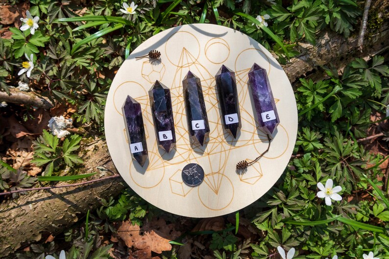 May include: Five amethyst points, labeled A through E, are arranged in a circle on a wooden board with a geometric pattern. The board is surrounded by green foliage.