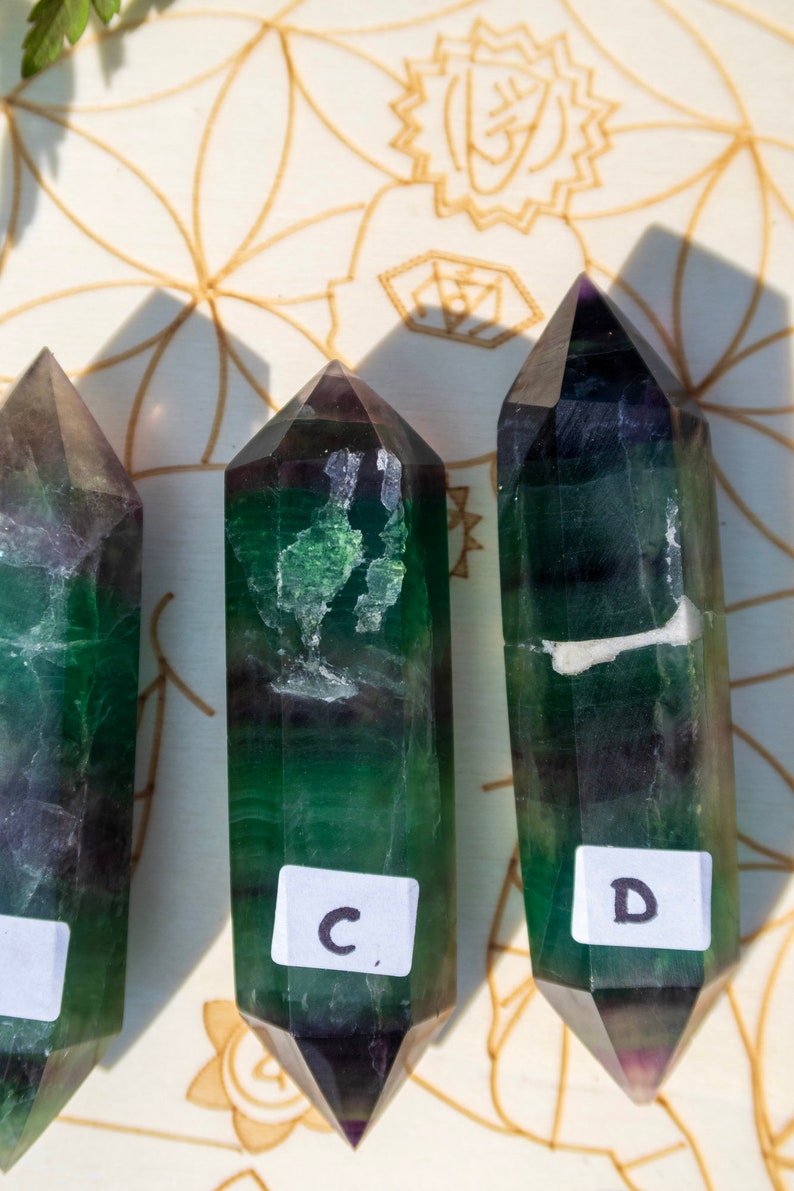 May include: Three green and purple fluorite crystal points with labels "C", "D", and a blank label. The crystals are arranged in a row on a white surface with a brown pattern.