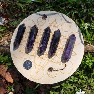 May include: Six purple amethyst points arranged in a circle on a wooden board with a geometric design. The board is surrounded by green foliage.