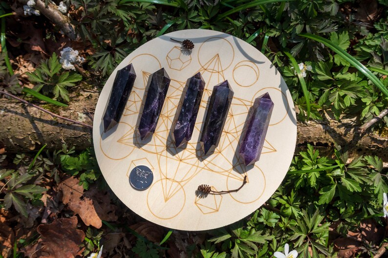 May include: Six purple amethyst points arranged in a circle on a wooden board with a geometric design. The board is surrounded by green foliage.