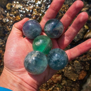 Puede incluir: Cinco esferas de fluorita verde y azul, tambi&eacute;n conocidas como bolas de cristal, se sostienen en la mano de una persona. Las esferas son lisas y pulidas, con una variedad de tonos de verde y azul.