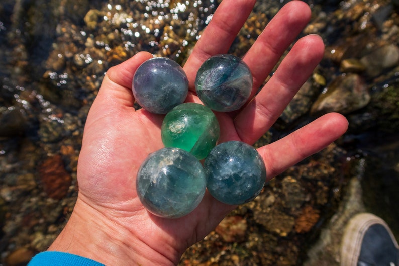 Puede incluir: Cinco esferas de fluorita verde y azul, tambi&eacute;n conocidas como bolas de cristal, se sostienen en la mano de una persona. Las esferas son lisas y pulidas, con una variedad de tonos de verde y azul.