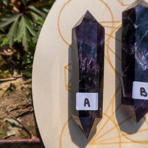 May include: Two dark purple amethyst crystal points on a wooden board with a geometric design. The points are labeled "A" and "B".