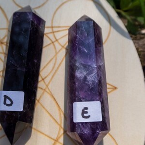 May include: Two purple amethyst crystal wands on a wooden surface with a geometric pattern. The wands are labeled with the letters "D" and "E".