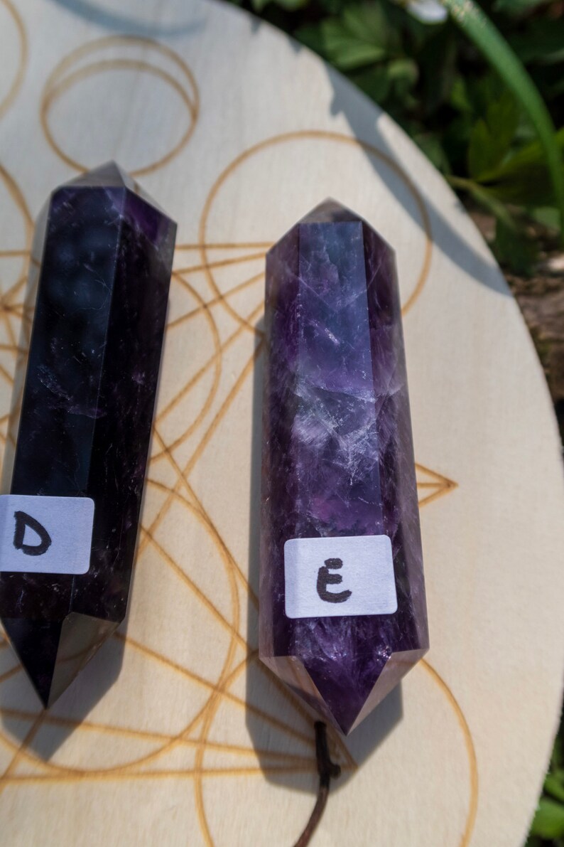 May include: Two purple amethyst crystal wands on a wooden surface with a geometric pattern. The wands are labeled with the letters "D" and "E".