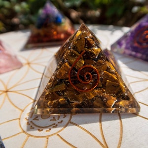 May include: A clear pyramid-shaped crystal with a copper wire spiral inside. The crystal is filled with small, golden-brown stones. The pyramid is sitting on a white surface with a geometric pattern.