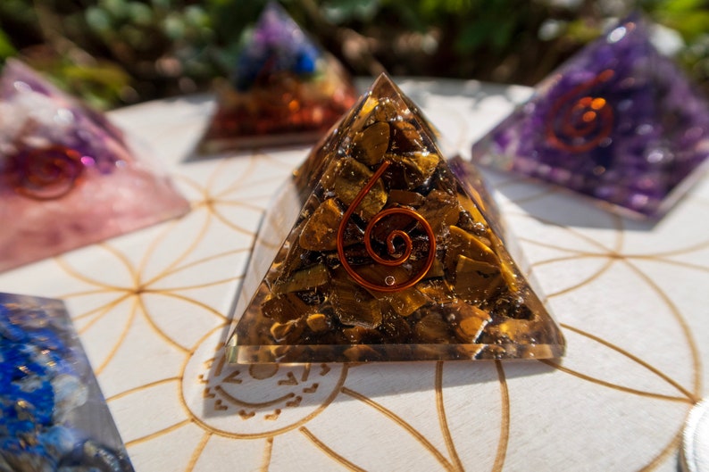 May include: A clear pyramid-shaped crystal with a copper wire spiral inside. The crystal is filled with small, golden-brown stones. The pyramid is sitting on a white surface with a geometric pattern.
