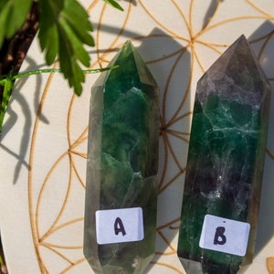 May include: Two green fluorite crystal points, labeled A and B, are displayed on a wooden surface with a flower of life pattern. The crystals are a deep green with some darker green and black inclusions.