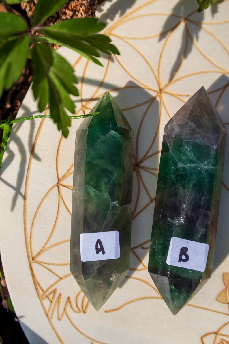 May include: Two green fluorite crystal points, labeled A and B, are displayed on a wooden surface with a flower of life pattern. The crystals are a deep green with some darker green and black inclusions.