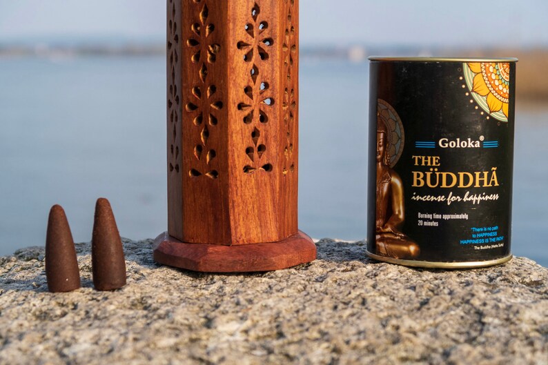 May include: Two brown incense cones next to a wooden incense burner and a black tin of Goloka The Buddha incense for happiness. The tin has a gold Buddha image and text that says "Burning time approximately 20 minutes".