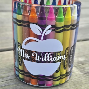 May include: A clear, cylindrical pencil holder filled with colourful crayons. The holder features a white apple design with the text "Mrs. Williams" in cursive. The crayons are arranged vertically, showcasing a spectrum of colours.
