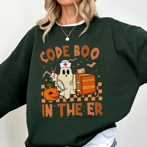 May include: Dark green sweatshirt with a Halloween-themed graphic. The design features a ghost wearing a nurse's hat, a pumpkin, and the text "CODE BOO IN THE ER". The graphic also includes a medical cart, syringe, and bats.