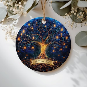 Puede incluir: Un adorno redondo de cerámica con un fondo azul oscuro y un árbol dorado brillante. Las ramas del árbol tienen pequeñas luces de color naranja intenso. Una cinta dorada en la base del tronco muestra el texto Allison 2024 en letra negra.