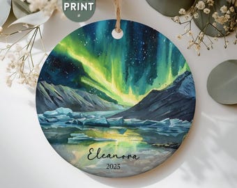 May include: Round Christmas ornament with a watercolor painting of the aurora borealis over a mountain range and a lake. The ornament has a two-sided print and includes the name "Eleanora" and the year "2025".