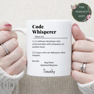 May include: White ceramic mug with a black and white definition of "Code Whisperer" with the name Timothy at the bottom. The mug is held by a person's hands.