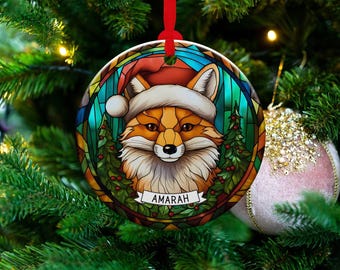 May include: A round Christmas ornament featuring a fox wearing a Santa hat. The fox is surrounded by a stained-glass design with Christmas trees and berries. The name "AMARAH" is displayed on a banner.