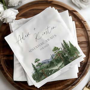 May include: White wedding napkins with a watercolor illustration of a forest scene. The text reads "Adam + Kirsten Adventure Awaits 13.11.2024".